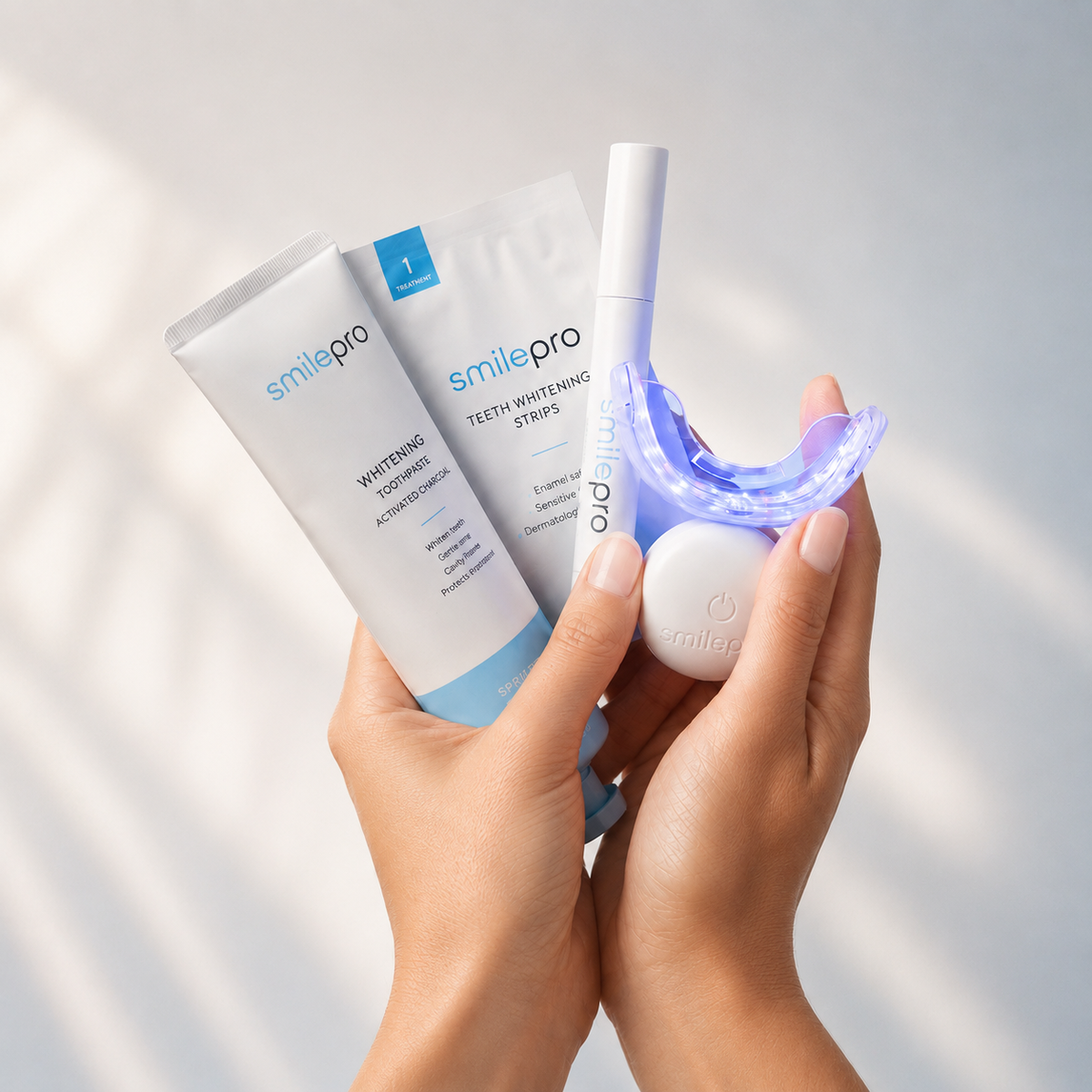 Hands holding SmilePro whitening toothpaste, teeth whitening strips, a whitening pen and the LED light